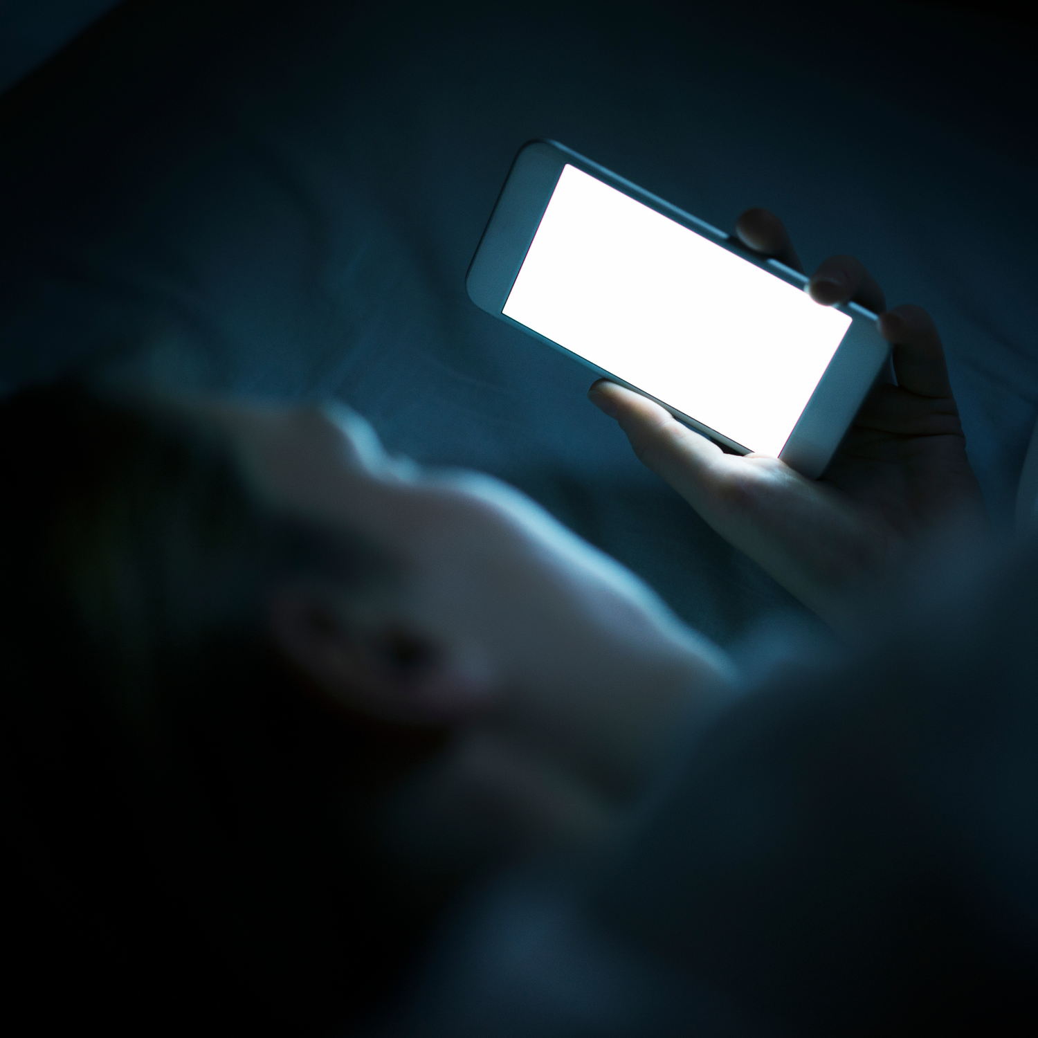 Persoon ligt in bed en kijkt naar een fel verlicht scherm van een smartphone - blog over de impact van blauw licht op slaap en gezondheid